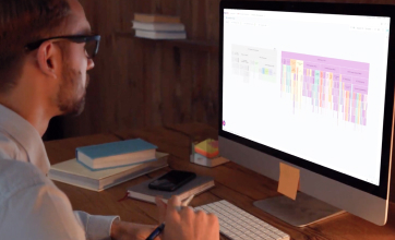 Image of a young man wearing glasses, focused on analysing data on a computer screen which displays colourful bar graphs.